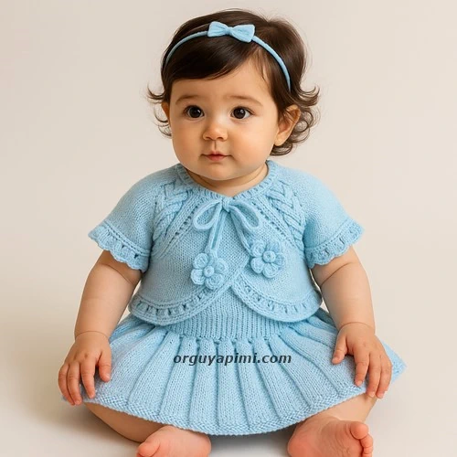 Açık mavi örgü kız bebek takımı – pileli fırfırlı etek ve burgulu bolero modelini giymiş 1 yaşında oturan bebek.